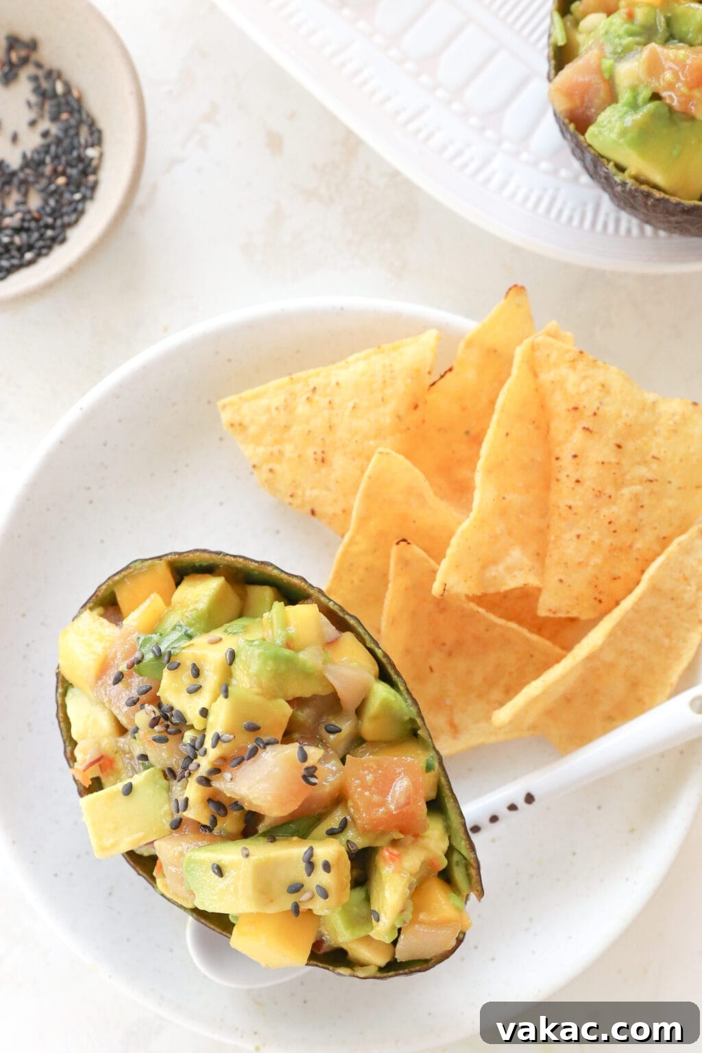 An avocado skin filled with cubed avocado, tuna and mango habanero salsa is on a white plate. There is a white spoon under the avocado and beside it are some chips.