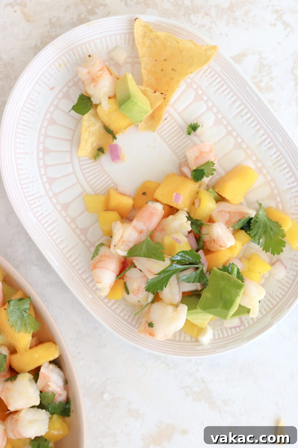 Speedy Prawn Ceviche with Vibrant Mango Avocado 6 A white plate with delicate pink details holds a generous serving of Prawn, Mango, and Avocado salad, accompanied by a handful of crisp tortilla chips. In the background, a larger platter of the same vibrant salad is visible, highlighting the red onion, chopped shrimp, and cubed mango components.
