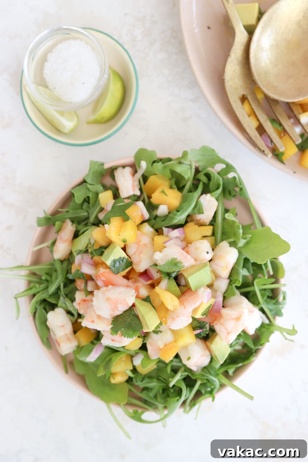 Speedy Prawn Ceviche with Vibrant Mango Avocado 8 A pink ceramic plate holds a generous serving of Prawn, Mango, and Avocado salad, artfully arranged on a bed of fresh arugula. In the softly blurred background, a large platter of the same vibrant salad is visible, along with two elegant gold serving spoons, setting a inviting and abundant scene.