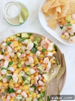 An overhead shot of a salad on a pink serving plate with gold serving spoon and fork. The salad has chopped shrimp, cubed mango, cubed avocado, and cilantro in it. In the corner is a glass of soda water with lime and a plate of tortilla chips with some of the salad on it.