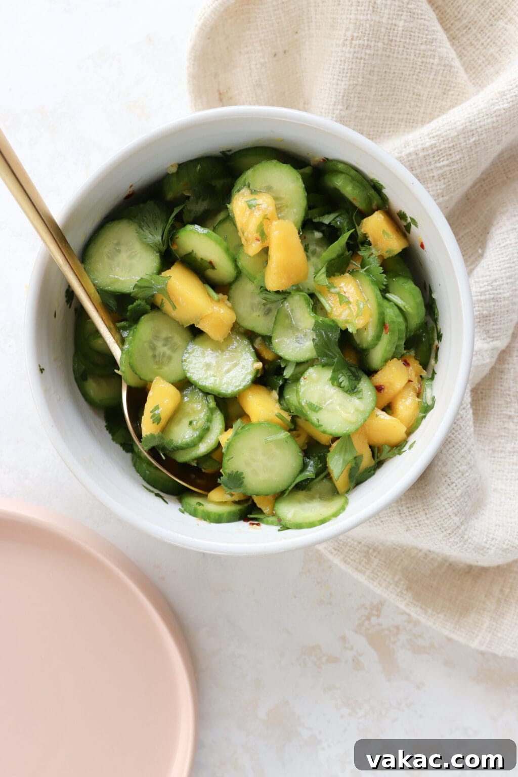 Easy Quick Pickled Mango Recipe 11 A pristine white bowl is filled with a refreshing cucumber mango salad, featuring vibrant greens and a gold fork poised for eating. A soft pink plate is visible to the left, adding a pop of color to the scene.