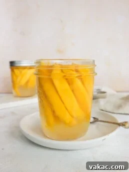A mason jar filled with mangoes is on a white plate. There is a spoon to the right of the plate. In the background is another mason jar with pickled mangoes.