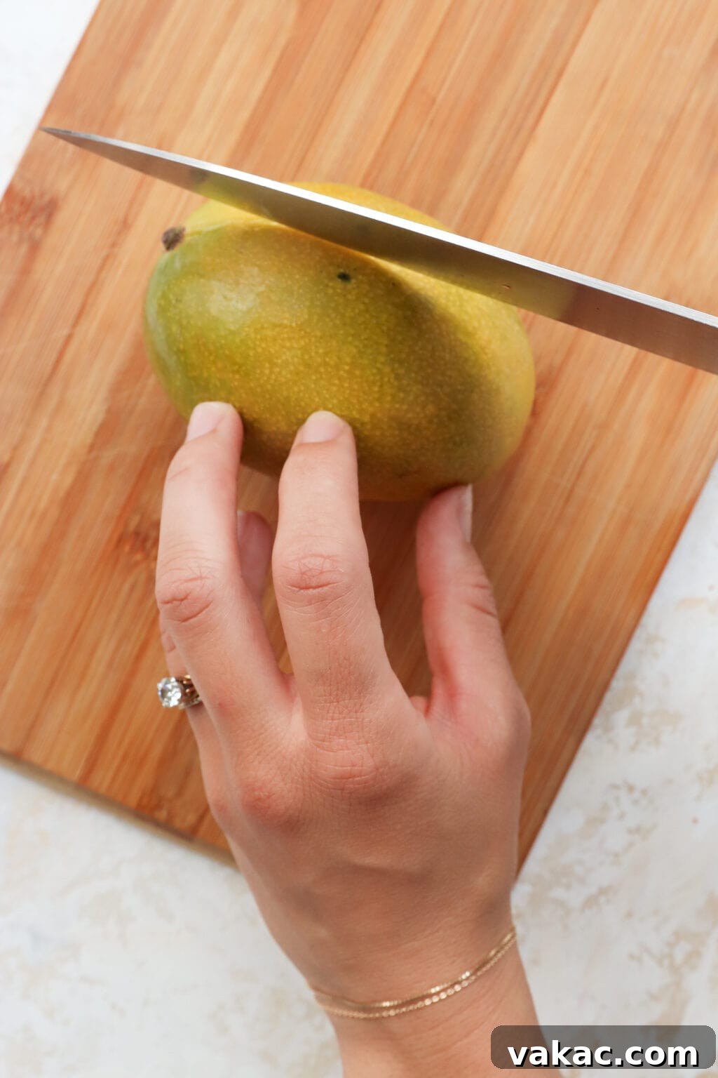 Easy Quick Pickled Mango Recipe 4 A mango with shades of green and yellow rests on a wooden cutting board, being held by a woman's hand as a chef's knife begins to slice into it.