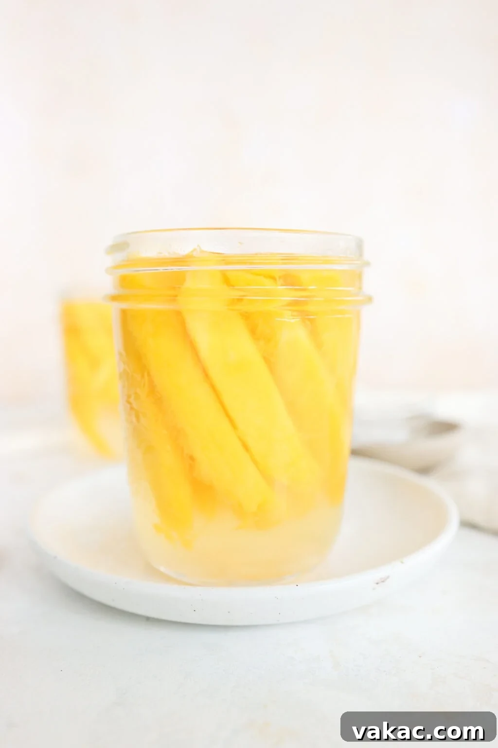 Easy Quick Pickled Mango Recipe 10 A pristine mason jar, brimming with golden pickled mangoes, is elegantly placed on a white plate with a spoon resting beside it. Another jar of pickled mangoes is visible in the soft-focus background.