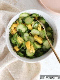 A white bowl is filled with sliced cucumber, diced mangoes, and cilantro, served with a gold spoon.