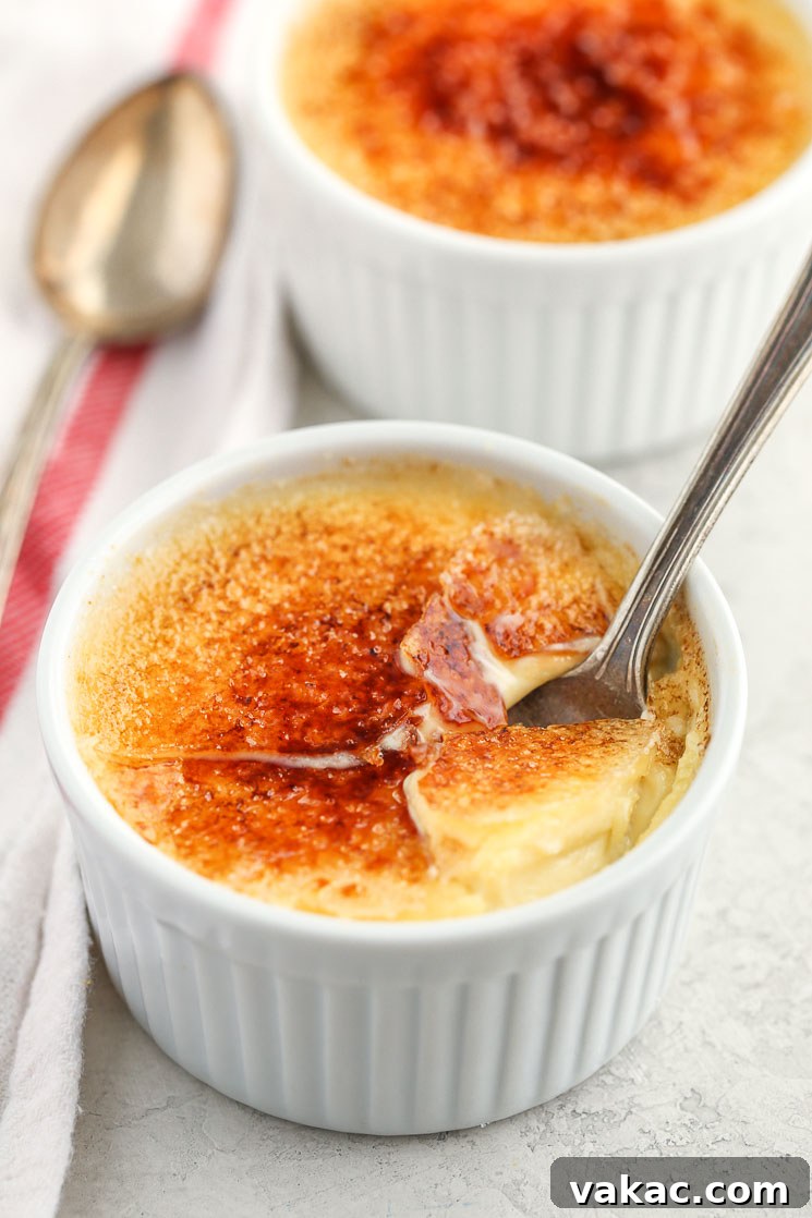 Crème Brûlée Made Simple 6 A close-up of a white ramekin filled with crème brûlée and a spoon inside of it along with another crème brûlée and white napkin, showing the beautiful texture and topping.