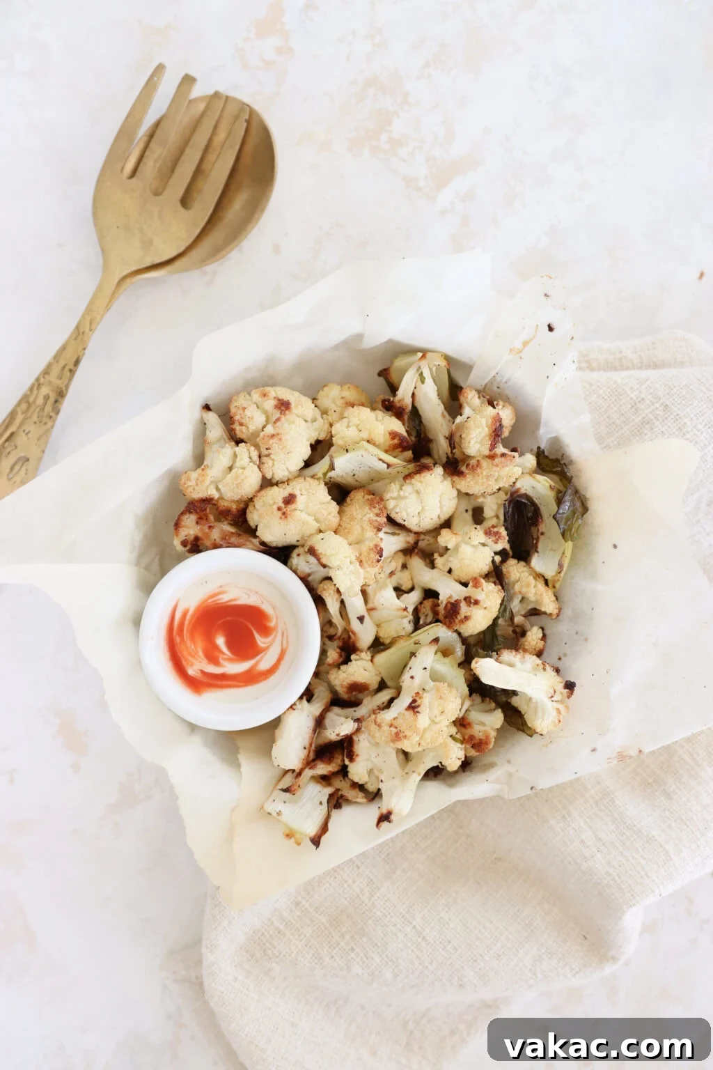 Meal Prep Magic Healthy Roasted Cauliflower Made Easy 7 A bowl lined with parchment paper holds roasted cauliflower florets. There is a small ramekin filled with white yogurt and a swirl of hot sauce. There is a set of gold serving spoons beside the cauliflower bowl.