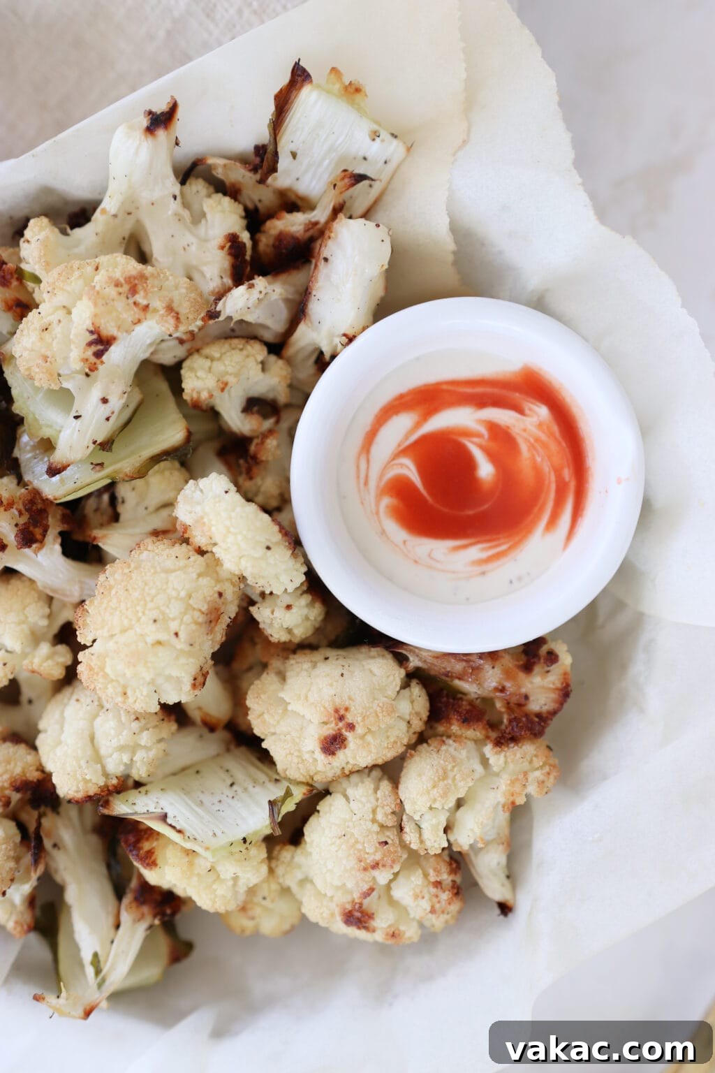 Meal Prep Magic Healthy Roasted Cauliflower Made Easy 8 A bowl lined with parchment paper holds roasted cauliflower florets. There is a small ramekin filled with white yogurt and a swirl of hot sauce.