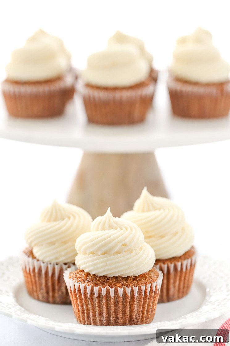 Spiced Carrot Cupcakes 5 A decorative white plate holding three carrot cake cupcakes topped with cream cheese frosting in front of a marble cake stand.