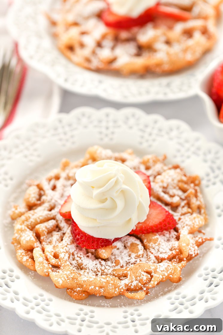 Two funnel cakes topped with powdered sugar, strawberries, and homemade whipped cream sitting on decorative white plates.