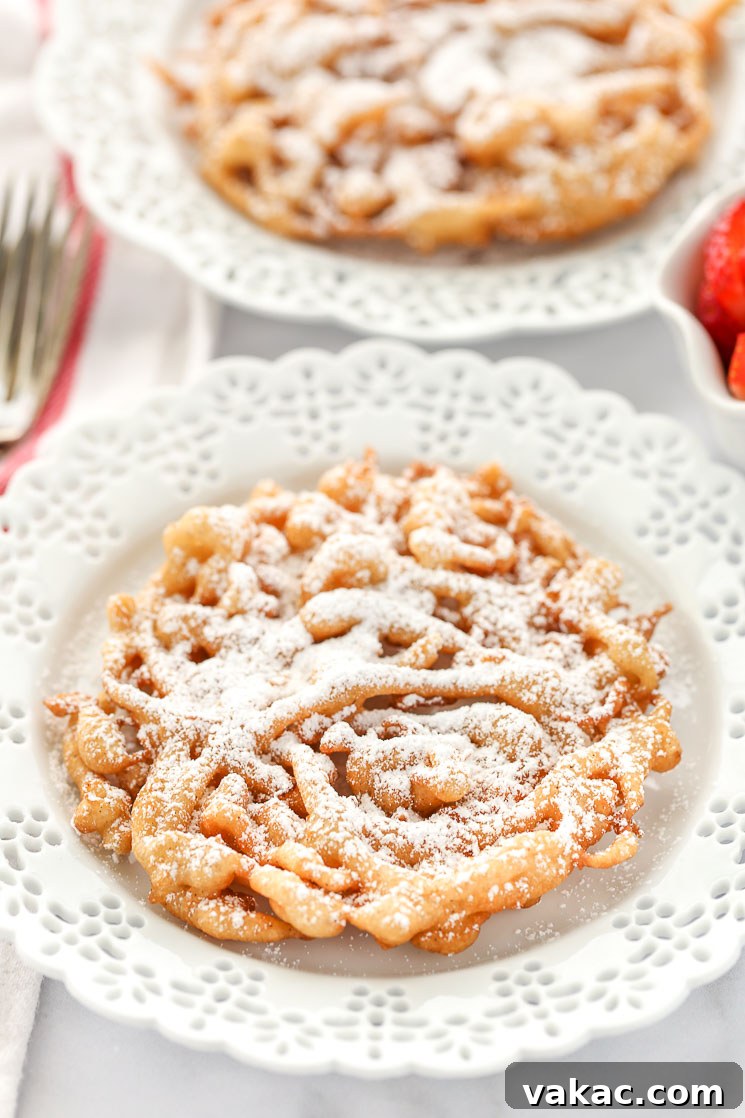 Two homemade funnel cakes topped with powdered sugar on decorative white plates and strawberries on the side.