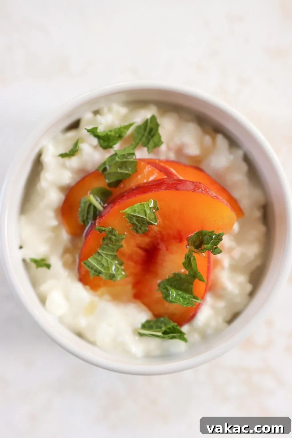 A white bowl is on an off white cloth. In the bowl is cottage cheese with slices of peaches topped with mint. There is a gold spoon beside the bowl.