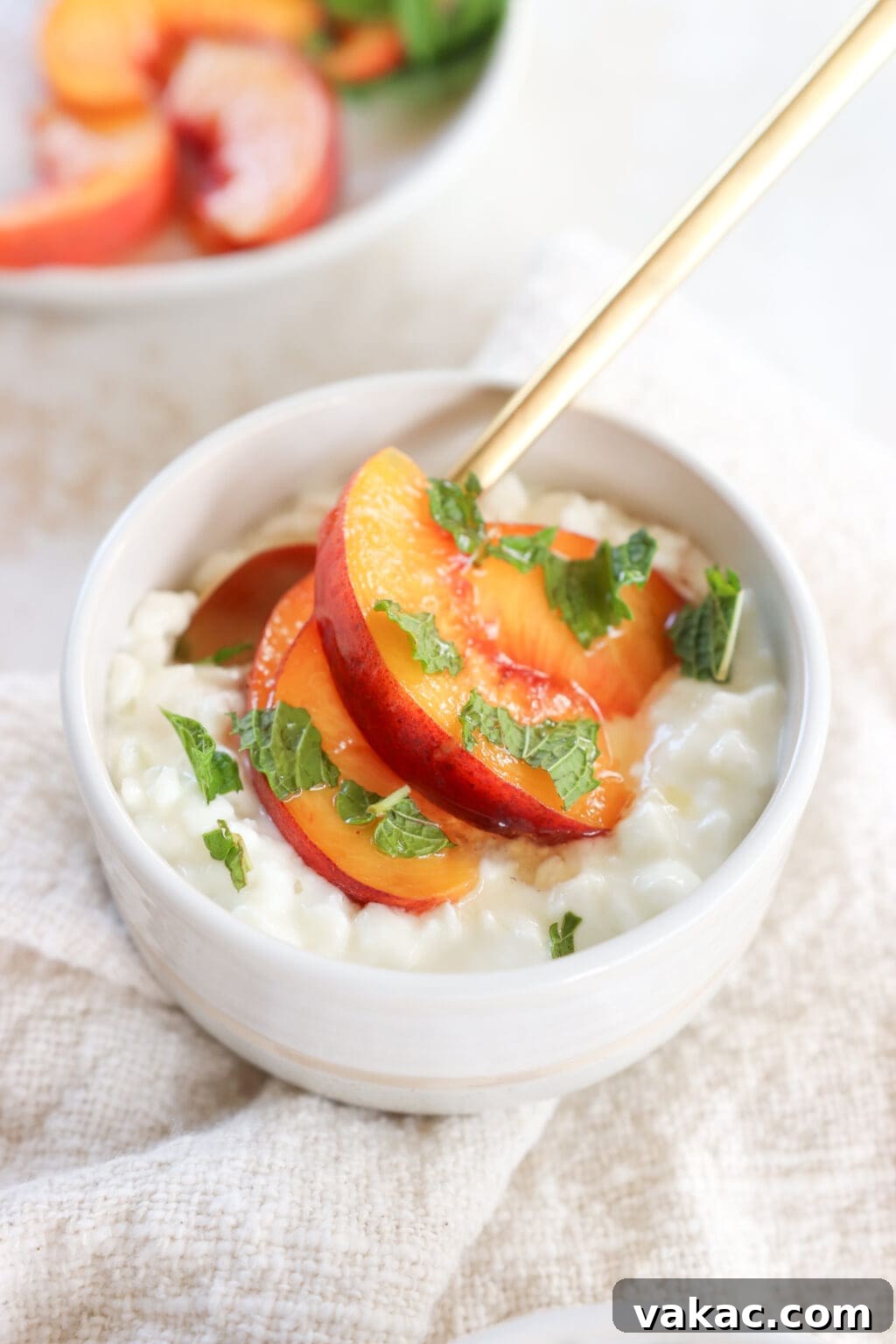 A white bowl is filled with cottage cheese, sliced peaches and topped with mint. There is a gold spoon in the bowl.