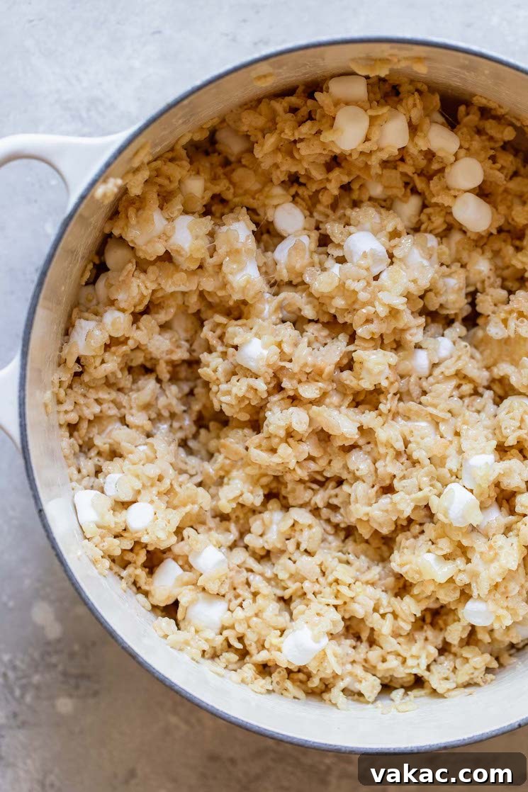 A Dutch oven with crispy rice cereal mixed into the melted butter and marshmallow mixture, creating a sticky, uniform base for the treats.