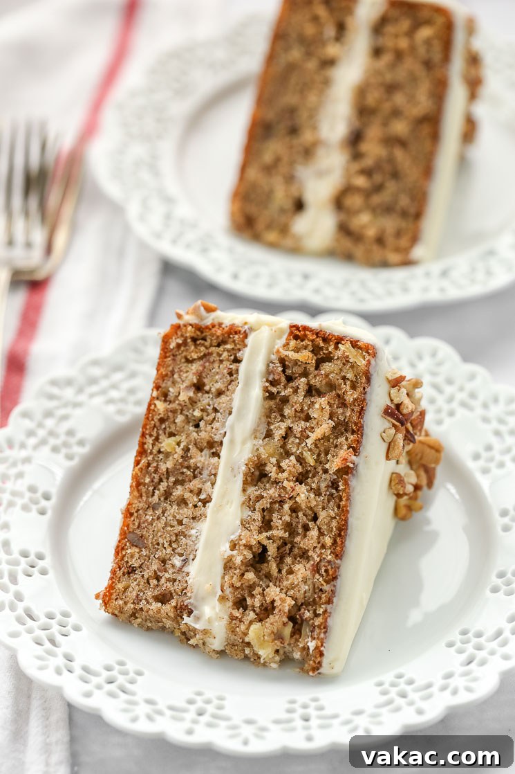 A beautifully presented slice of hummingbird cake on a decorative white plate, with another slice and a fork in the soft-focus background, highlighting its moist layers and creamy frosting.