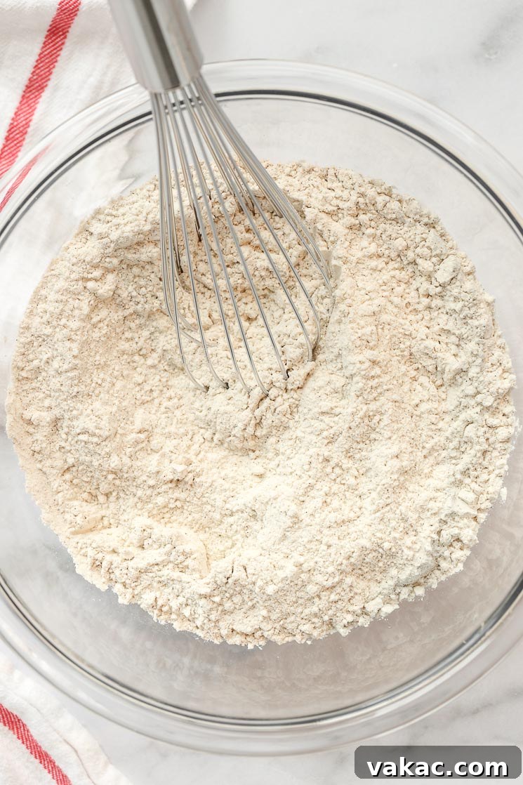 A clear glass bowl filled with the dry ingredients for a cake, featuring a whisk resting on the side, ready for mixing.