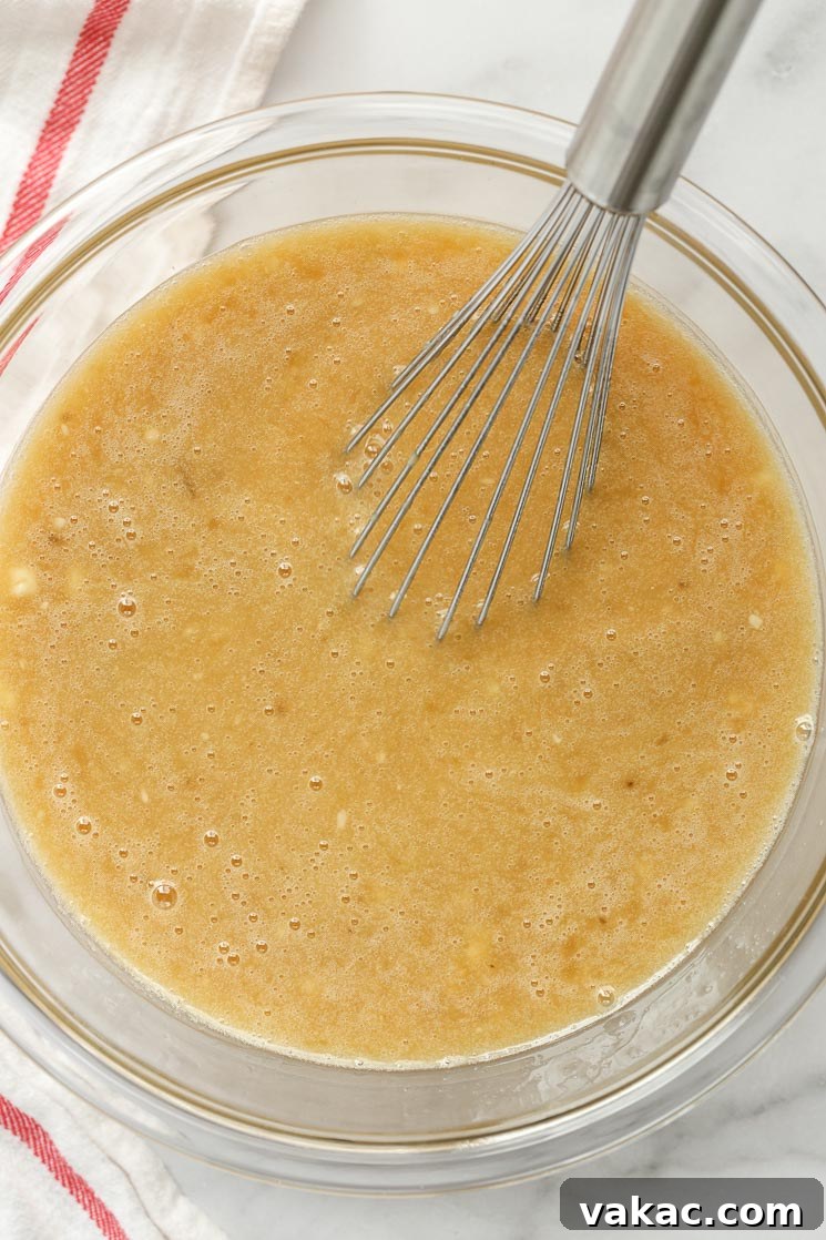 A clear glass bowl containing the liquid wet ingredients for a cake, with a metal whisk resting inside, illustrating the readiness for combining.