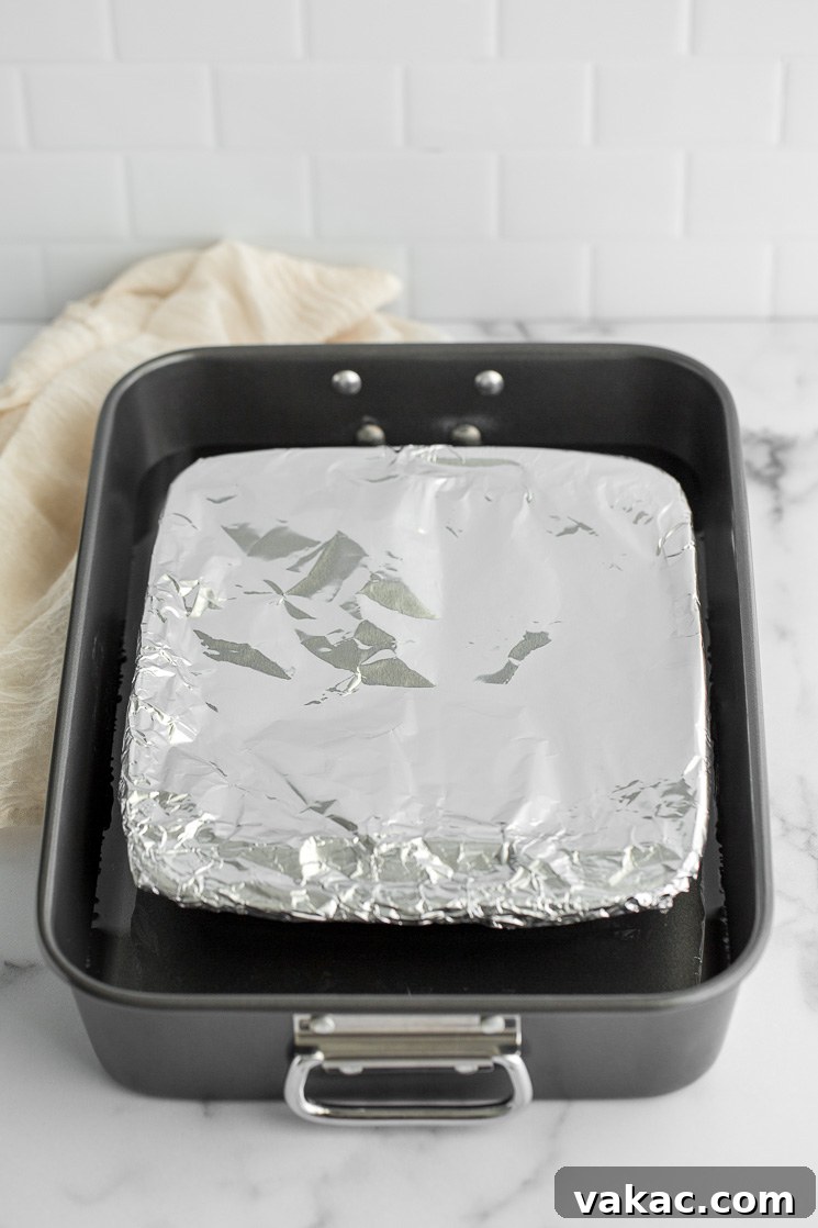 A glass baking dish filled with sweetened condensed milk, tightly covered with aluminum foil, resting inside a larger roasting pan, poised for the water bath method.