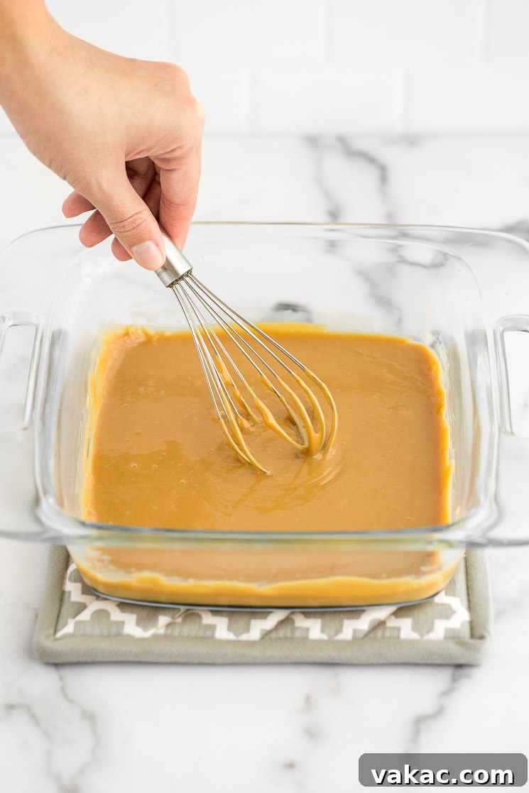 Freshly baked dulce de leche in a clear glass baking dish, being whisked vigorously to achieve a smooth, creamy consistency.