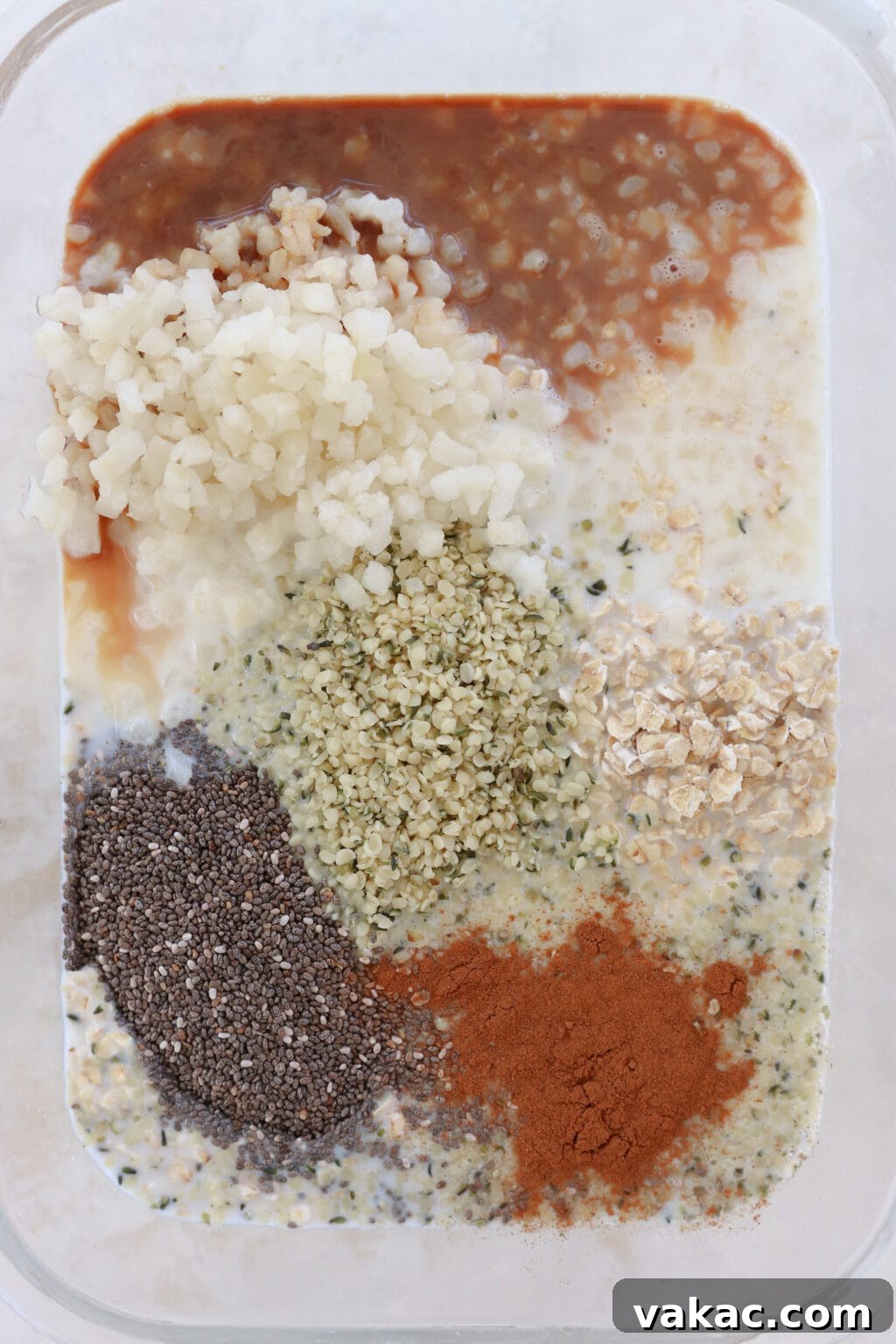 Make-Ahead Cinnamon Roll Overnight Oats 4 A glass container filled with unmixed ingredients for cinnamon roll overnight oats, showcasing quick oats, hemp hearts, chia seeds, vanilla extract, milk, maple syrup, salt, ground cinnamon, and cauliflower rice, ready to be combined.
