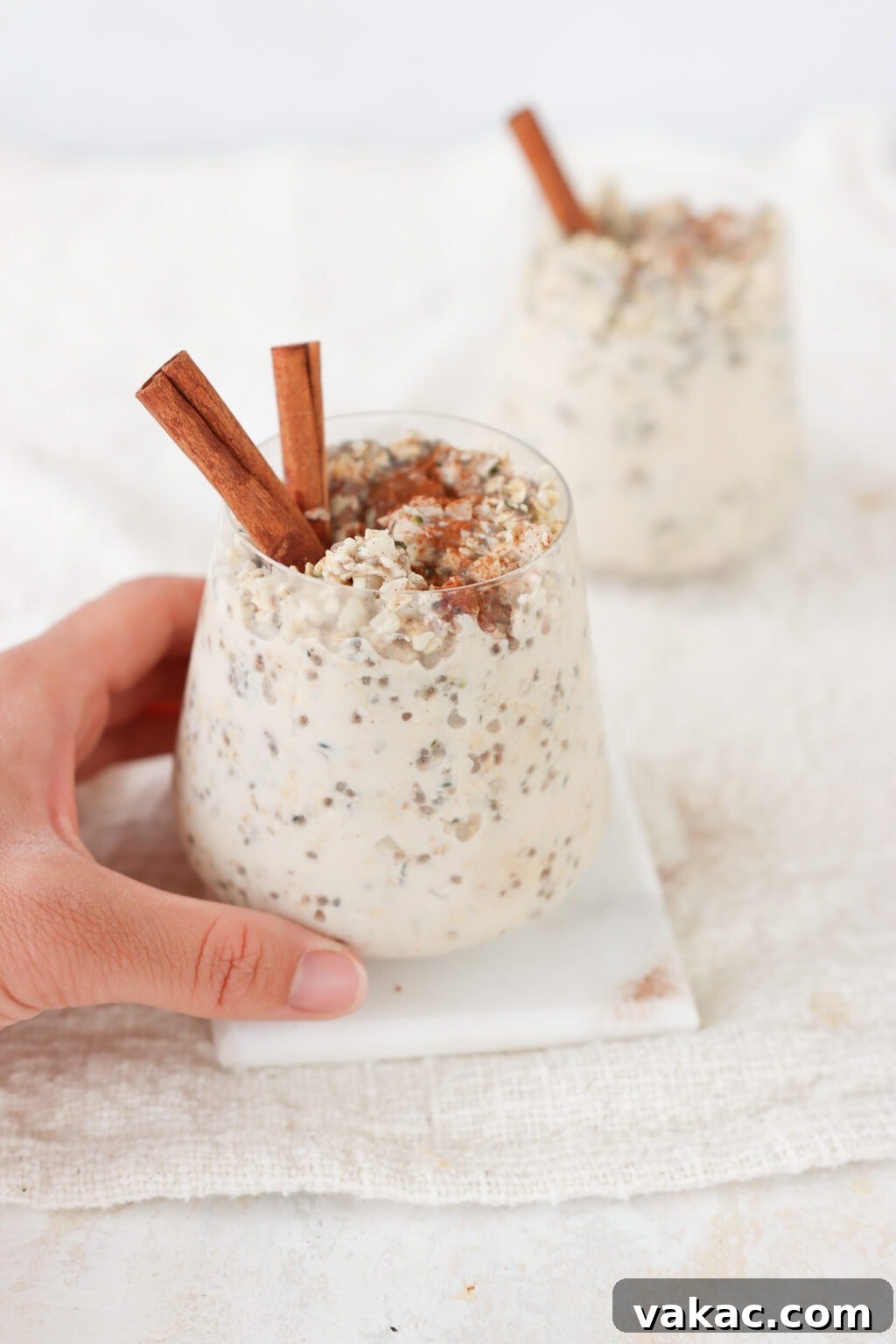 Make-Ahead Cinnamon Roll Overnight Oats 6 A hand gently reaching for one of two glasses filled with creamy cinnamon roll overnight oats, garnished with cinnamon sticks, positioned on a bright white surface, highlighting their ready-to-eat convenience.