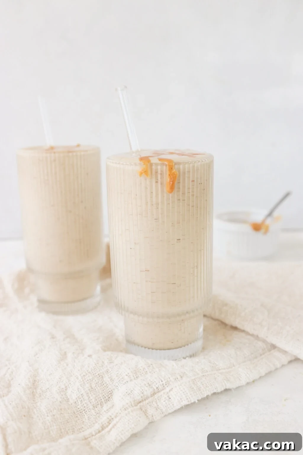 Two glasses filled with healthy peanut butter banana smoothie with a bowl of melted peanut butter on the side