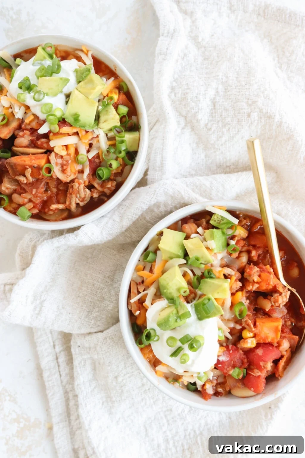 Comforting Slow Cooker Sweet Potato and Turkey Chili 6 Two white bowls of Slow Cooker Sweet Potato and Turkey Chili, generously topped with shredded cheese, avocado slices, and fresh cilantro, presented on a white tablecloth.
