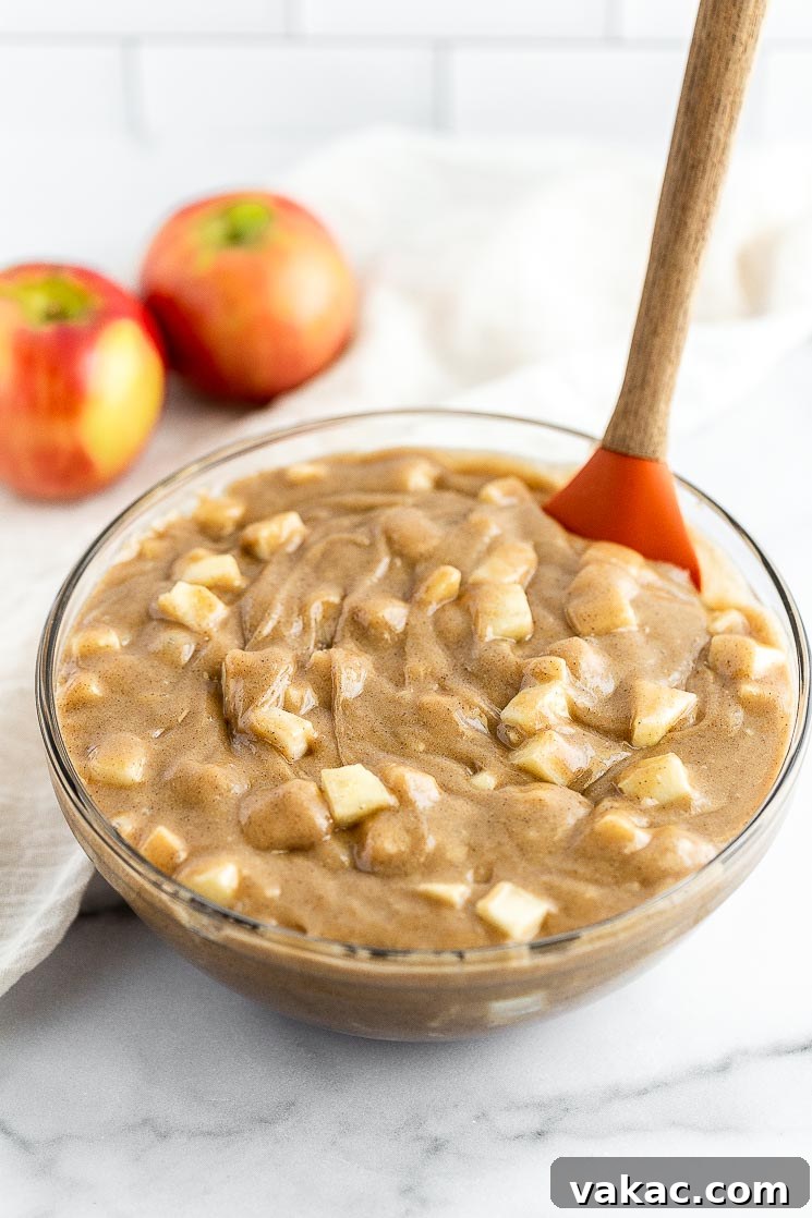 A close-up shot of a glass mixing bowl brimming with light brown cake batter, generously mixed with freshly chopped apples.