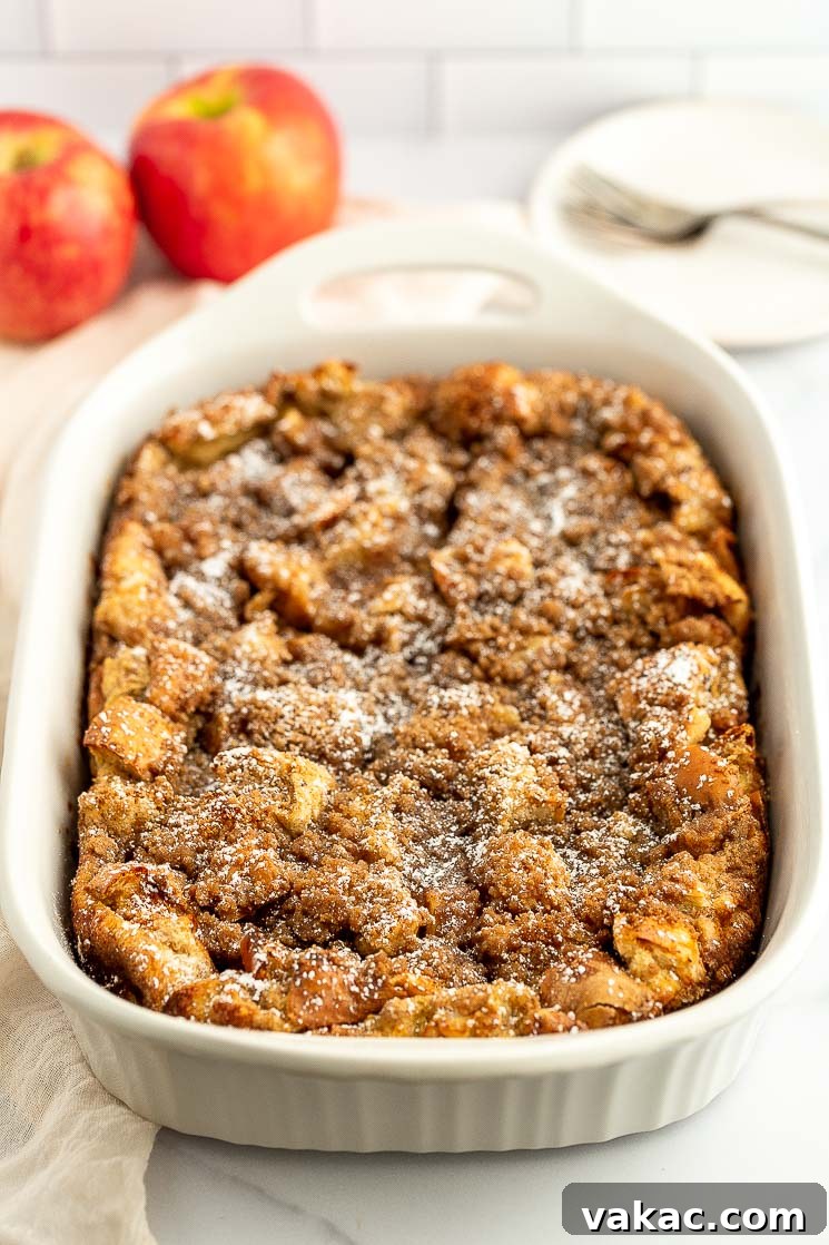 Cinnamon Apple Streusel French Toast Bake 2 Baked apple french toast casserole in a white baking dish, dusted with powdered sugar and surrounded by fresh apples.