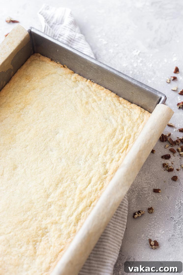 A baking pan lined with parchment paper, revealing a perfectly pre-baked shortbread crust, golden brown and ready for the pecan filling.