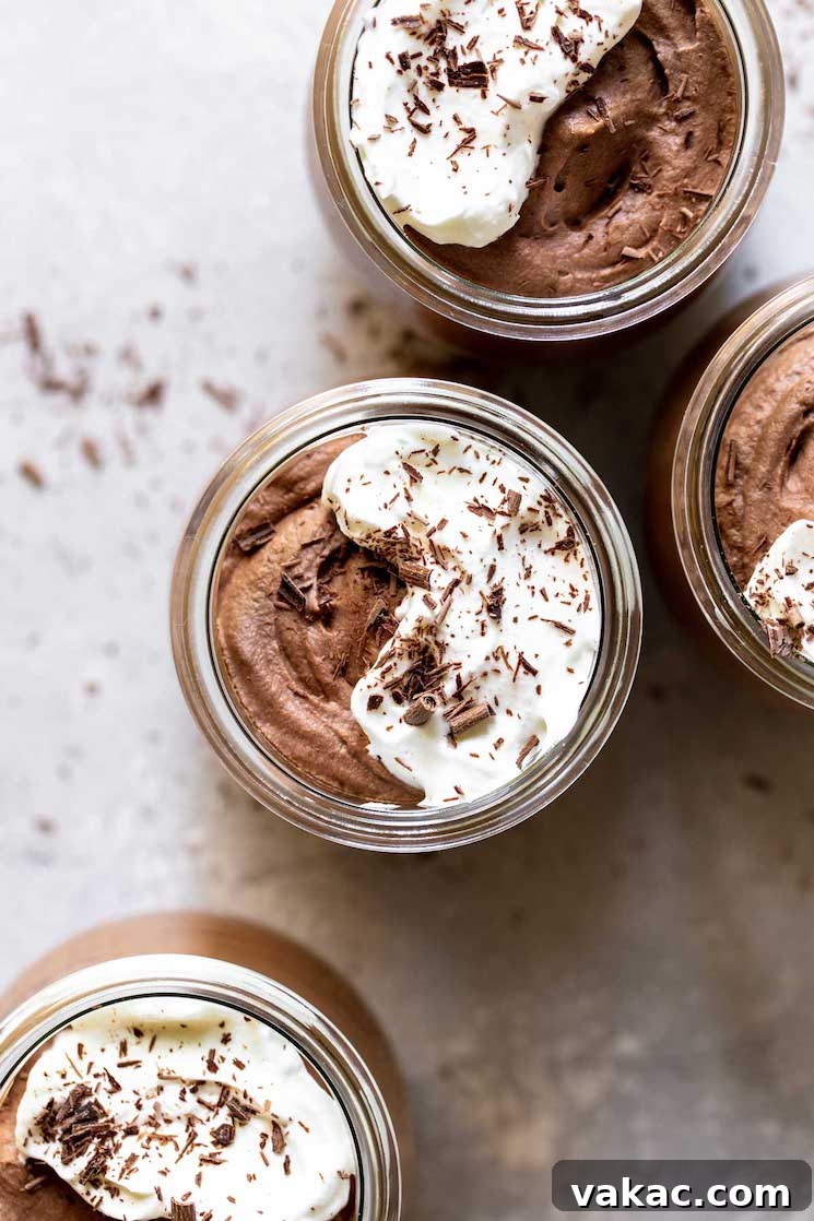 Velvet Chocolate Indulgence 7 Four small glass jars filled with chocolate mousse topped with whipped cream and chocolate shaving on a gray surface dusted with more chocolate shavings.