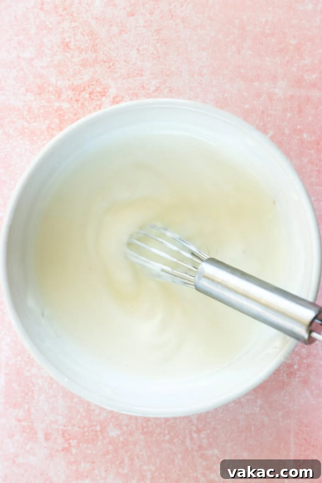 Just 4 Ingredients for Tangy Greek Yogurt Lime Crema 5 A close-up of Greek Yogurt Lime Crema being whisked to a smooth consistency in a white bowl with a metal whisk, on a pink countertop, showing the blending process.