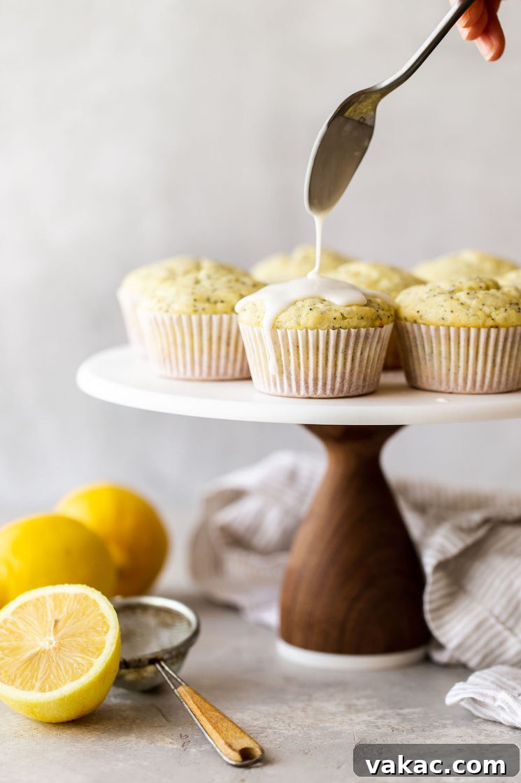 Zesty Lemon Poppy Seed Muffins 2 A beautifully arranged marble cake stand featuring golden Lemon Poppy Seed Muffins, some of which are being drizzled with a simple lemon glaze, highlighting their moist texture and vibrant appeal.