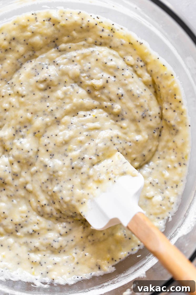 Zesty Lemon Poppy Seed Muffins 3 A clear glass bowl containing a vibrant yellow lemon poppy seed muffin batter, being gently mixed with a rubber spatula, showcasing its smooth consistency.