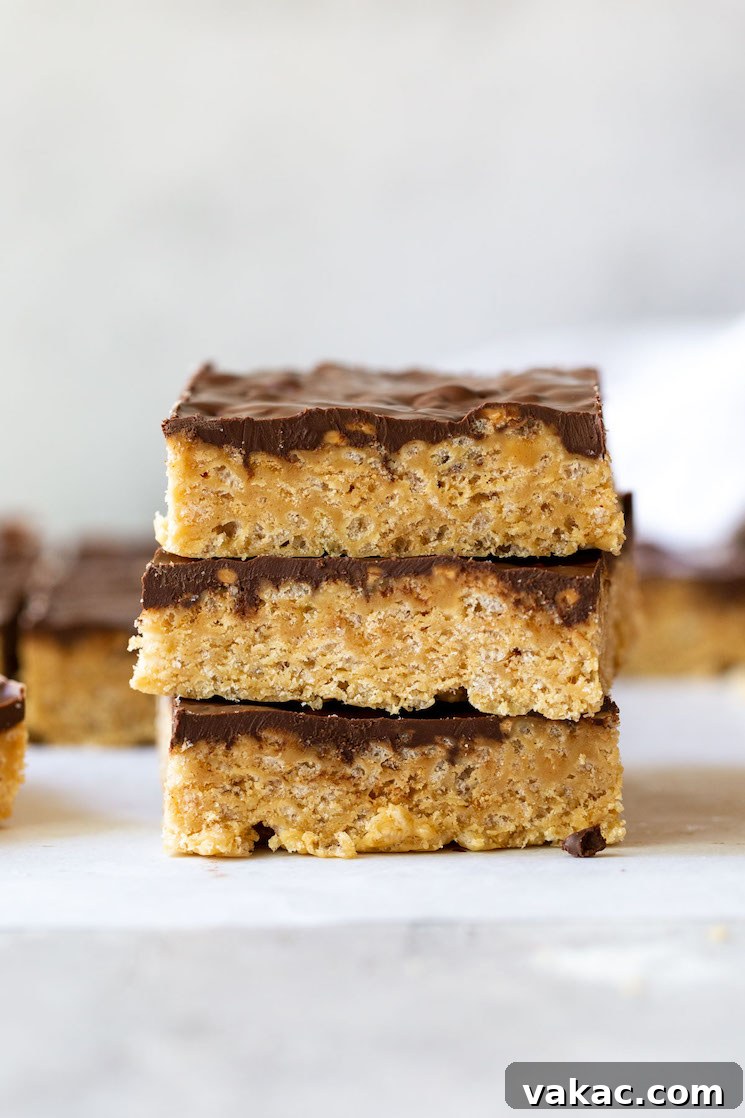 Peanut Butter Butterscotch Crispy Bars 2 A stack of three perfectly cut Scotcheroos on a rustic gray surface, with more bars blurred in the background, highlighting their delicious layers and appealing texture.
