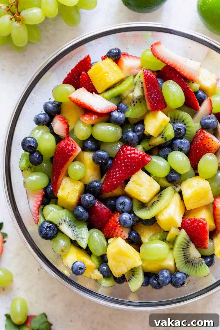 Sun Kissed Fruit Delight 5 A beautifully presented glass bowl overflowing with fresh fruit salad, strategically placed on a rustic gray surface, surrounded by individual fresh fruits like strawberries and lime halves.