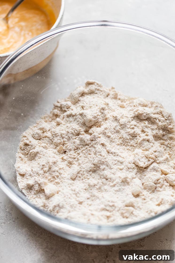 The dry ingredients for scones, including flour, sugar, and spices, are combined in a clear glass mixing bowl, with cold butter cut into small pieces amidst them.