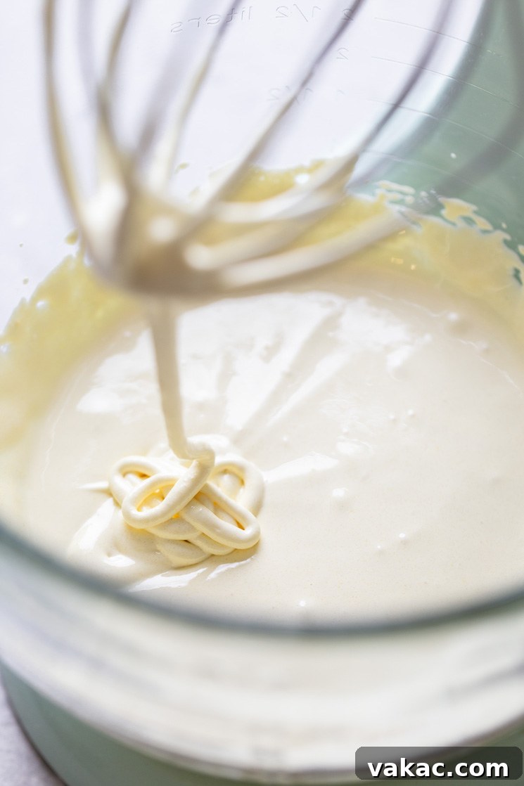 A close-up view of the smooth, pale yellow tres leches cake batter in a clear glass mixing bowl, with a whisk resting over the rim, hinting at the preparation process.