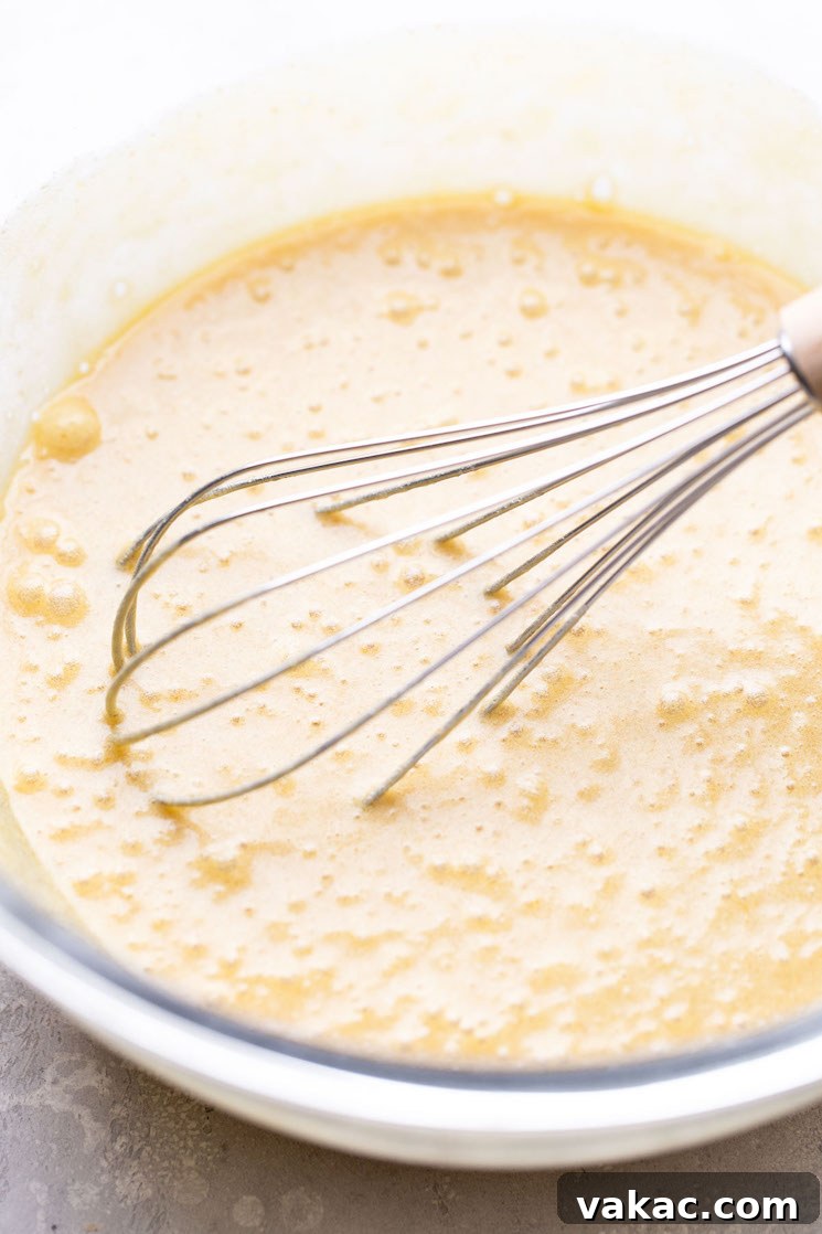 A clear glass mixing bowl filled with the glossy, amber-colored wet ingredients (melted butter, sugars, eggs, vanilla) being whisked smoothly together.