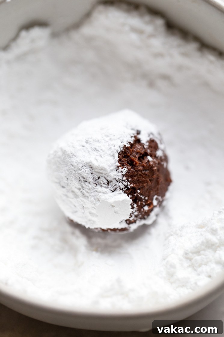 A single ball of dark chocolate cookie dough, perfectly shaped, being rolled in a bowl of fine powdered sugar, preparing it for baking.