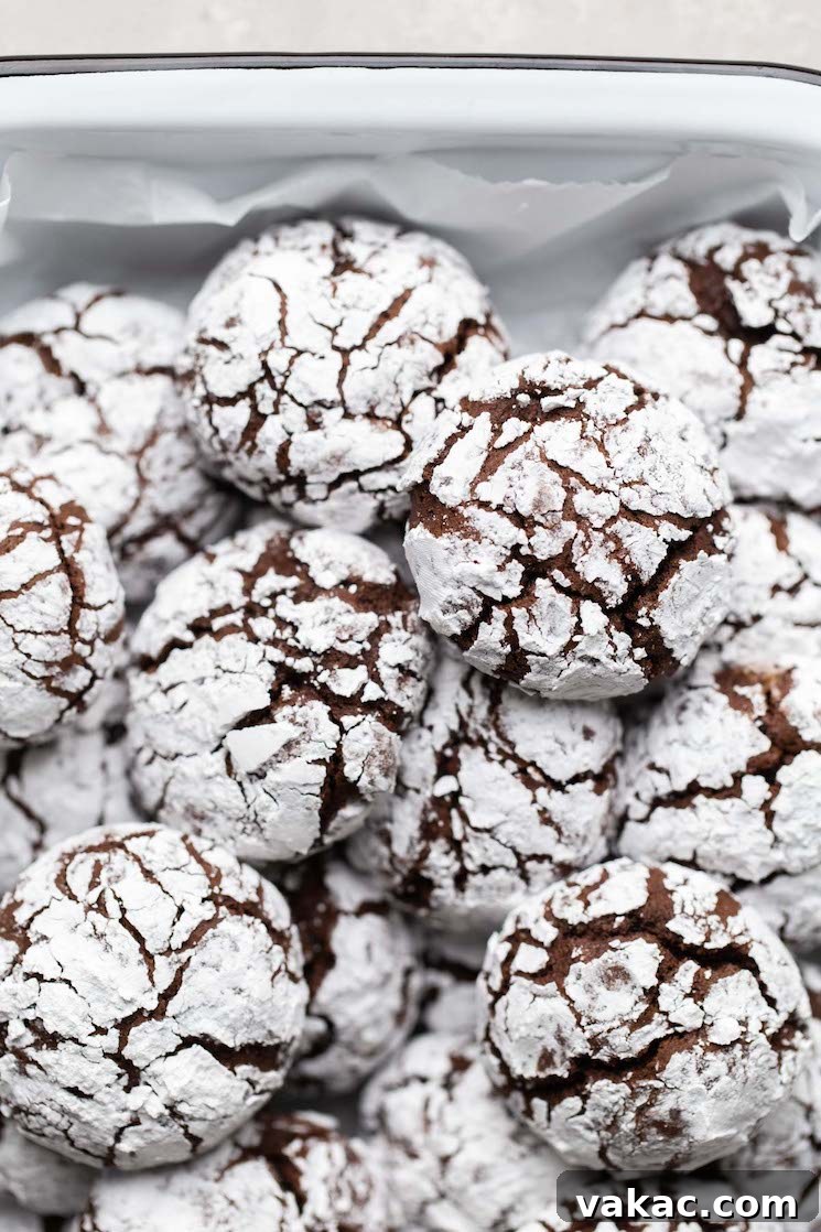 Several soft, fudgy chocolate crinkle cookies, perfectly dusted with powdered sugar, are stacked neatly in a white serving dish, inviting a delicious treat.
