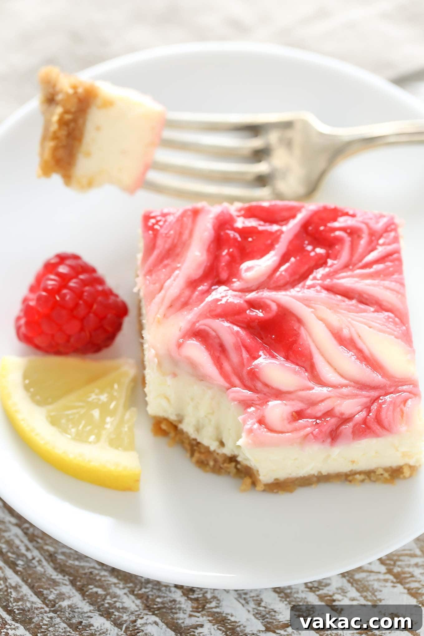 Zesty Lemon Raspberry Cheesecake Bars 4 A single, perfectly cut lemon raspberry cheesecake bar resting on a white plate, with a fork holding a small bite. A fresh lemon wedge and a raspberry garnish the plate.