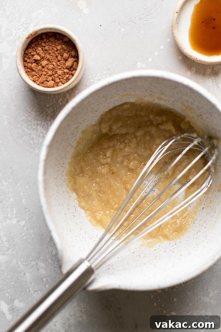 Melted butter and sugar in a mixing bowl with a whisk. A small dish of cocoa powder and vanilla extract sit on the side.