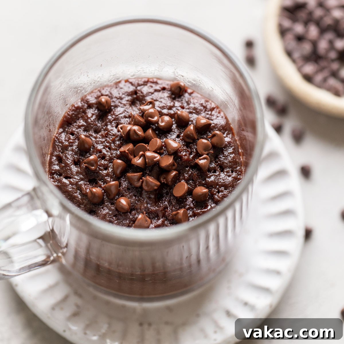 Microwaveable Mug Brownie Bliss 7 Overhead view of a microwave mug brownie in a glass mug. The mug sits on a white dessert plate, and a dish of mini chocolate chips rests on the side.