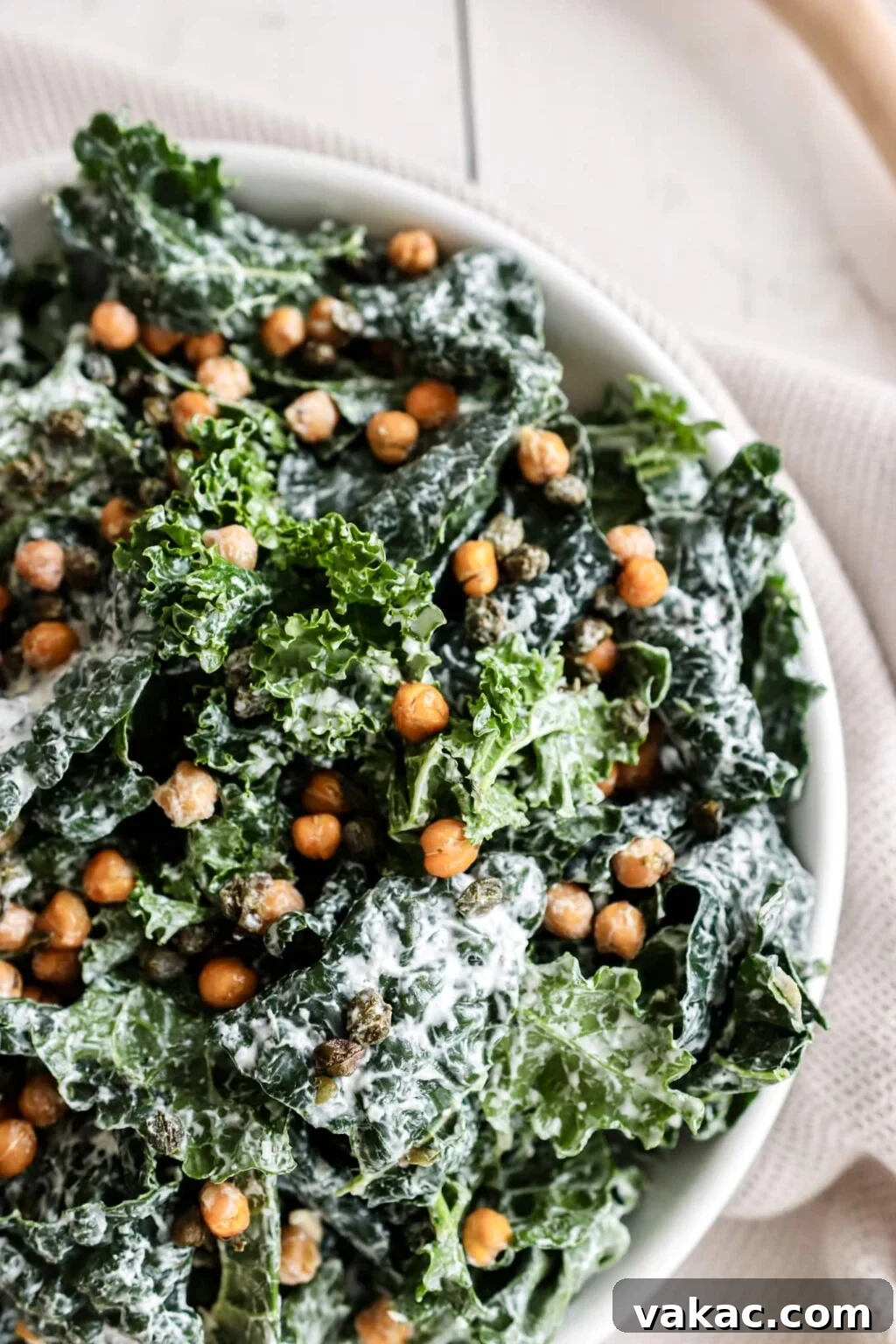 Chickpea kale caesar salad on a white plate over a neutral kitchen dish. Ingredients include: kale, chickpeas, capers, Greek yogurt, and garlic.