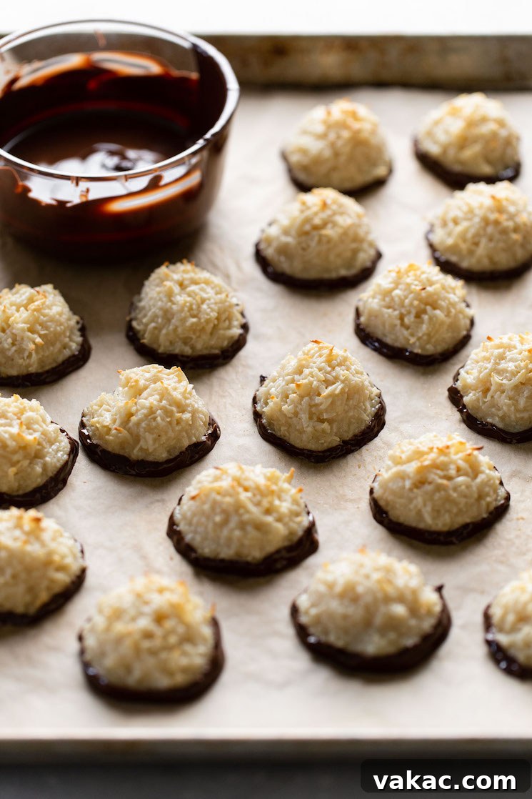 Chocolate dipped macaroons on a baking sheet. A dish of melted chocolate is also on the sheet.