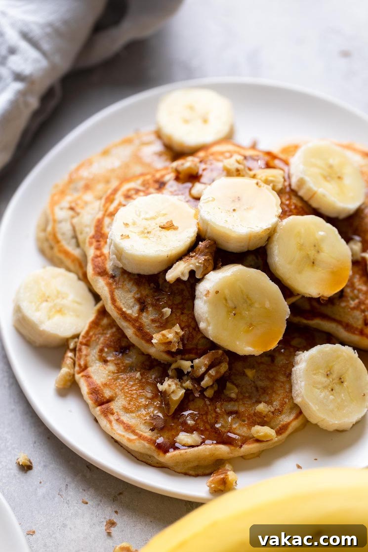 Four easy banana pancakes topped with syrup, banana slices, and walnuts on a white plate. A banana rests in the foreground.