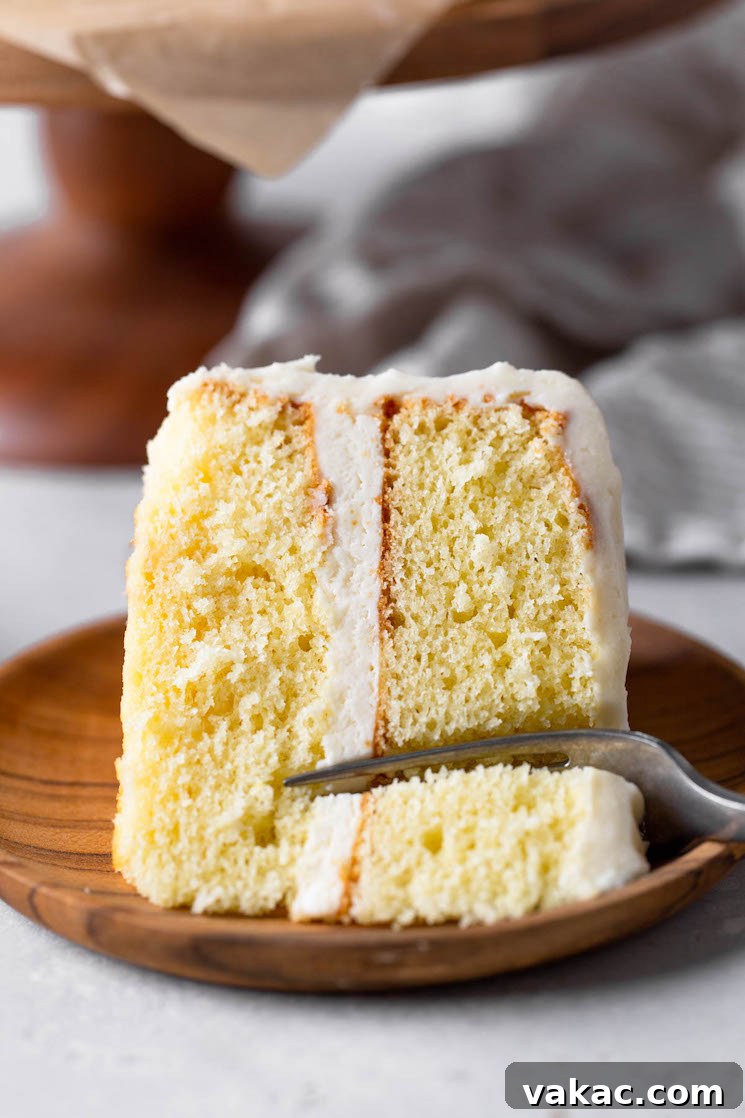 A delectable slice of fluffy vanilla cake, generously coated with creamy vanilla buttercream, laid invitingly on its side on a rustic wooden plate, with a fork poised to take the first delightful bite.
