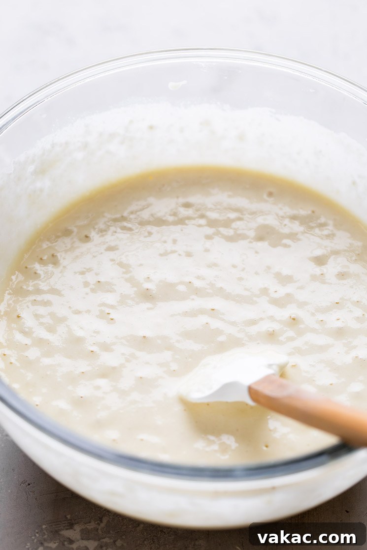 A glass mixing bowl full of waffle batter. A rubber spatula rests in the bowl.