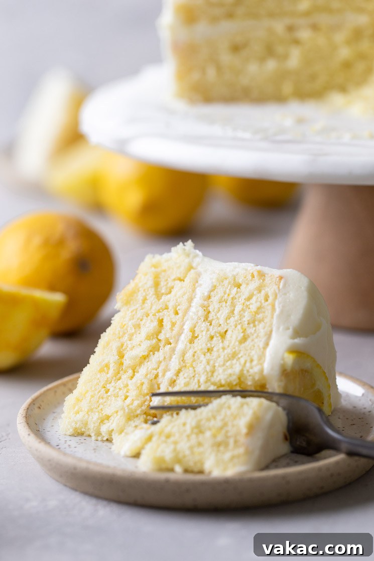 Sunshine Lemon Delight 9 A slice of homemade lemon cake lying on its side on a speckled plate. A fork is digging into the slice. The rest of the cake is on a cake stand in the background.